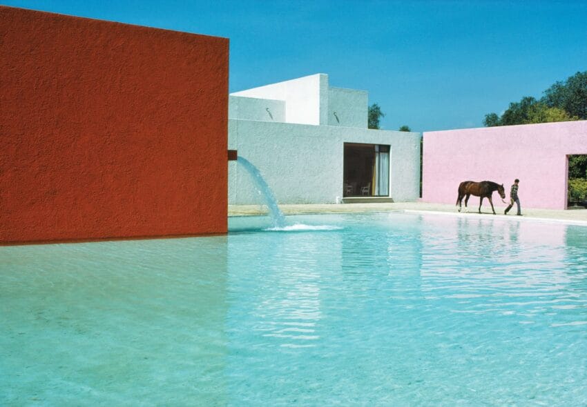 Modern architectural building with colorful walls surrounding a clear pool, a person walking a horse nearby under a blue sky.