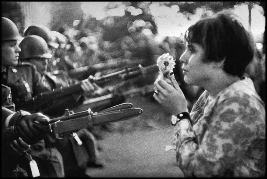 A woman holds a flower facing armed soldiers during a protest, capturing a moment of peaceful resistance.