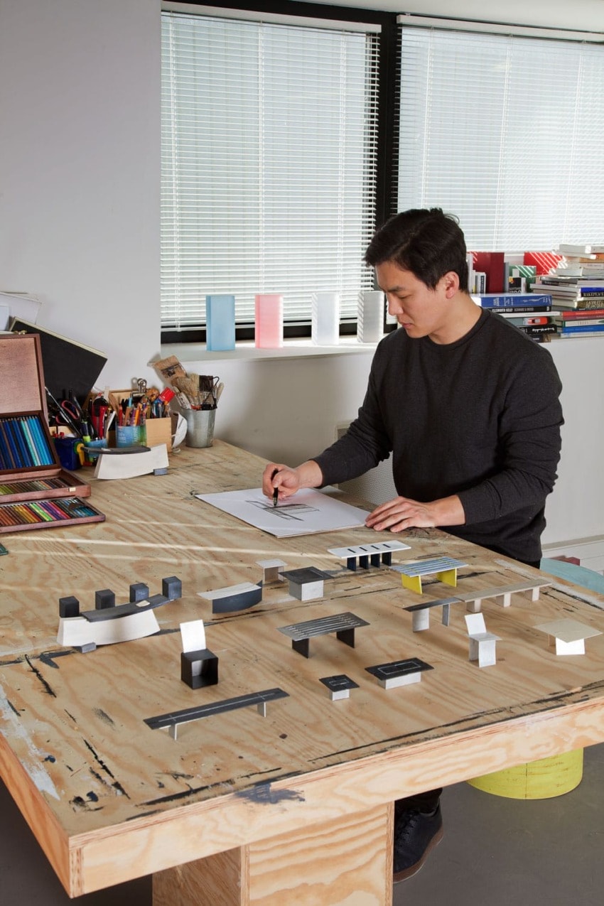 Architect working at a desk with models and sketches, surrounded by art supplies and books in a sunlit office.
