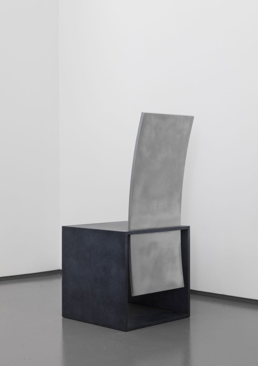 Modern minimalist metal chair with a sleek design placed in a corner against a white wall on a gray floor.