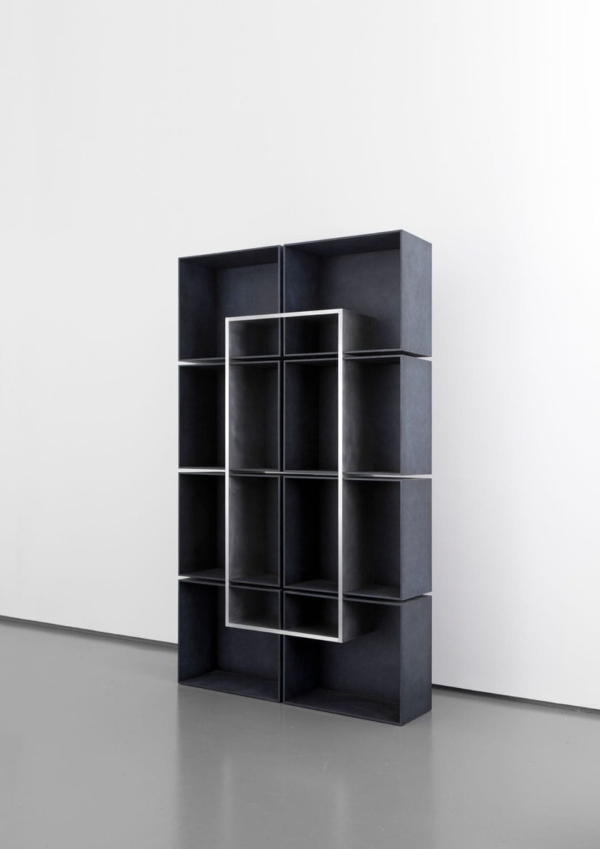 Modern black metal bookshelf with an abstract geometric design, standing against a plain white wall on a gray floor.