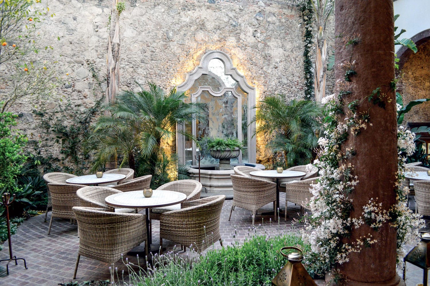 Outdoor courtyard with wicker chairs and tables, surrounded by lush greenery and a stone wall with an ornate fountain.