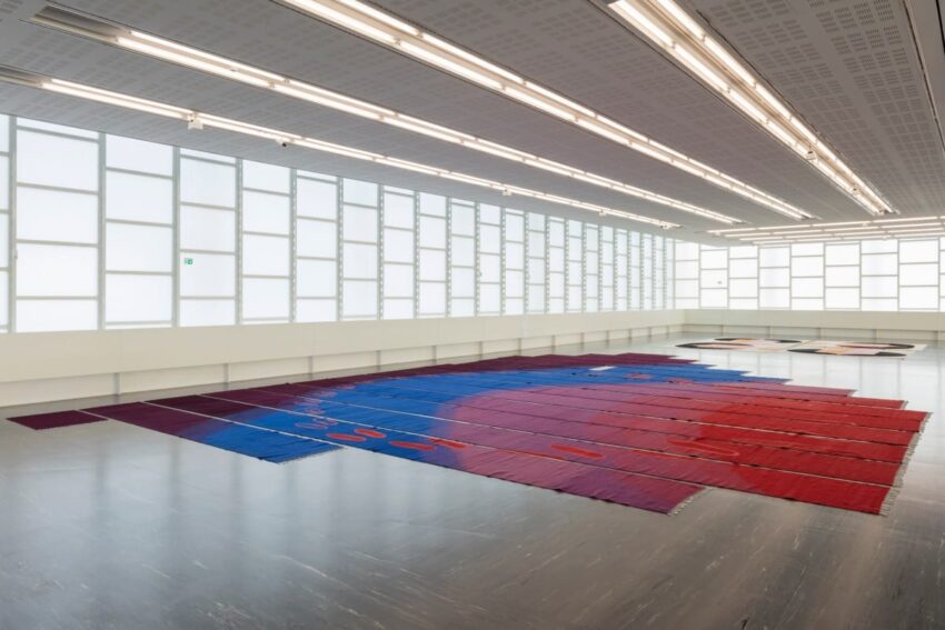 Colorful textile art installation on a gallery floor with large windows and overhead lights.