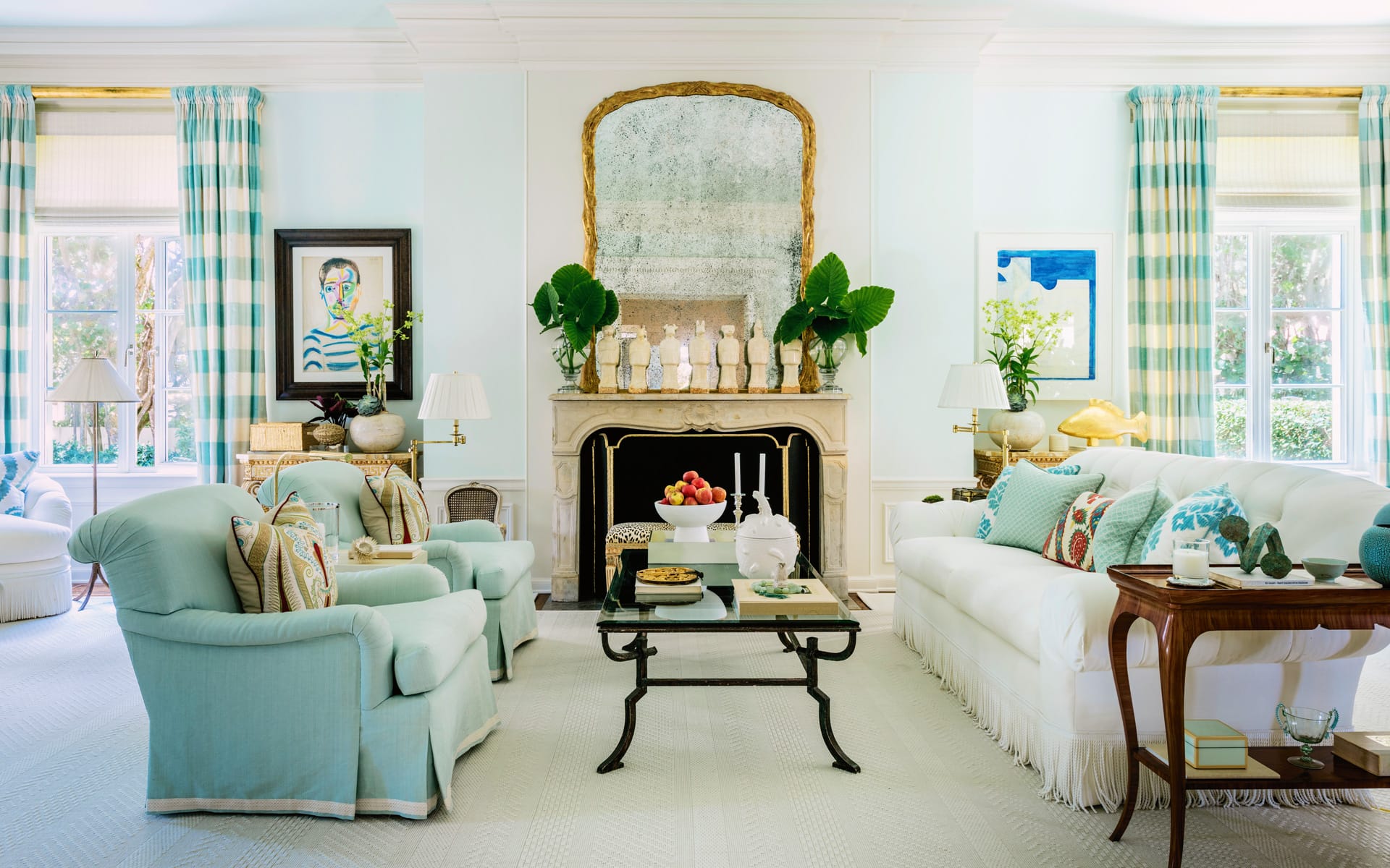 Elegant living room with blue chairs, white sofas, large mirror above the fireplace, plants, and checkered curtains.