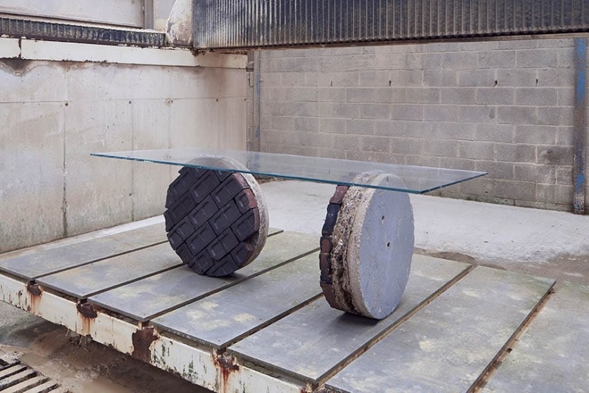 Glass table with industrial-style legs made from large concrete disks and brick elements in a minimalist, modern setting.