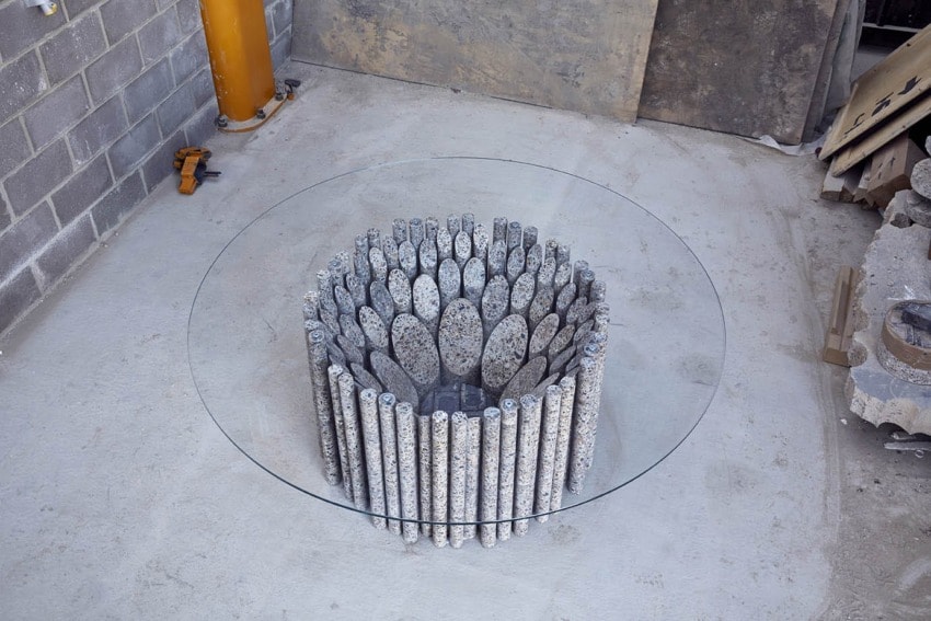 Round glass-top table with a base made of textured stone cylinders arranged in a circle on a concrete floor.