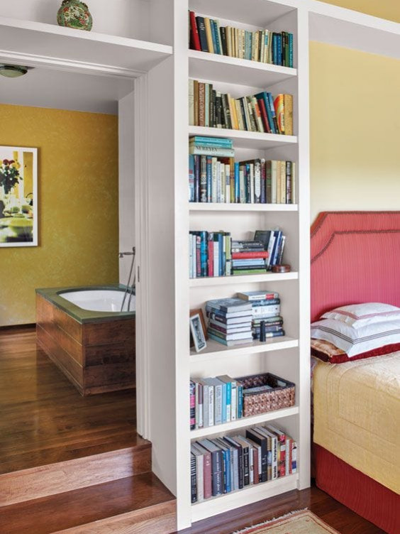 Bedroom with built-in bookshelf next to steps leading to bathroom with wooden floor and large bathtub.