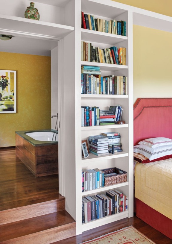 Bookshelf in a cozy bedroom with a red headboard and a view of a bathroom featuring a wooden bathtub.