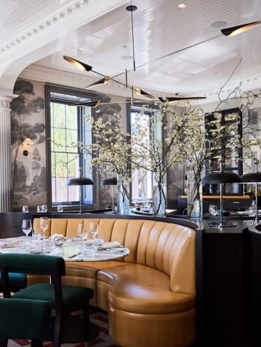 Elegant restaurant interior with a curved tan leather booth, dining table set with glassware, and decorative branches in vases.