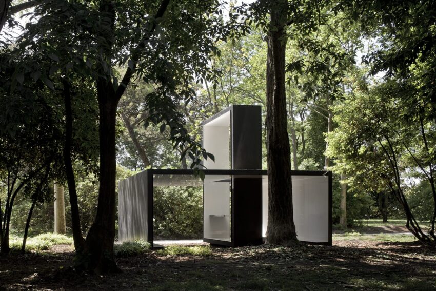 Modern glass and steel architectural structure in a forest setting surrounded by lush green trees and sunlight filtering through.