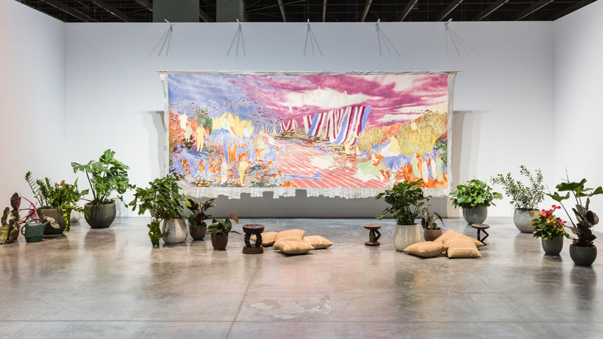 Colorful abstract tapestry on a gallery wall surrounded by various potted plants and cushions on a polished concrete floor.