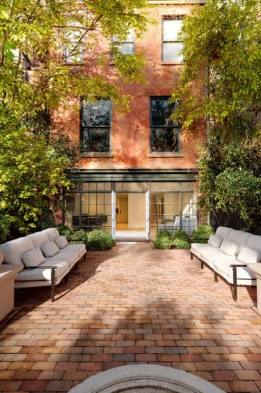 Brick patio with white cushioned benches, greenery, and large windows on a historic brick building in the background.