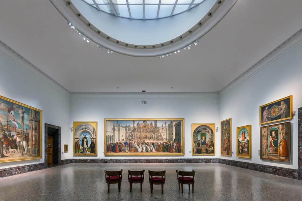 Interior of an art gallery showcasing large Renaissance paintings under a skylight, with four chairs facing the artworks.