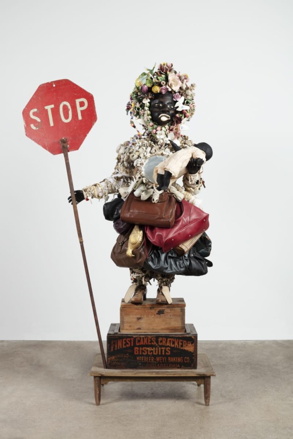 A sculpture of a figure with a floral headdress holding a stop sign and standing on a crate.