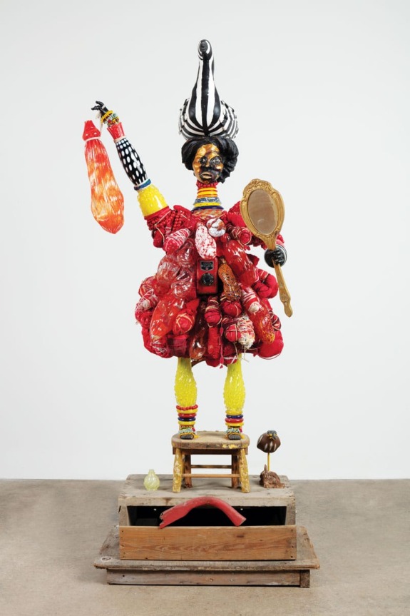 Colorful sculpture with striped hat, red elements, holding a mirror and a bottle, standing on stacked crates.