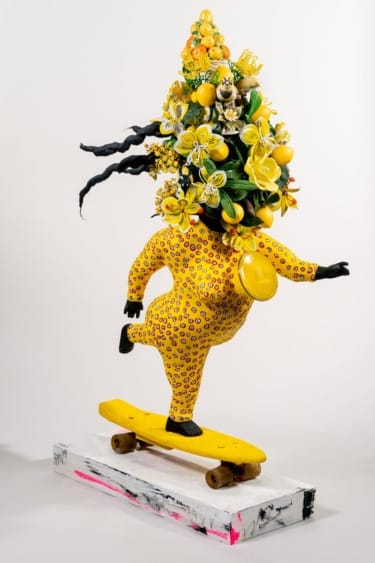 Statue of a colorful figure on a skateboard, wearing a floral headdress featuring vibrant yellow flowers and fruit.