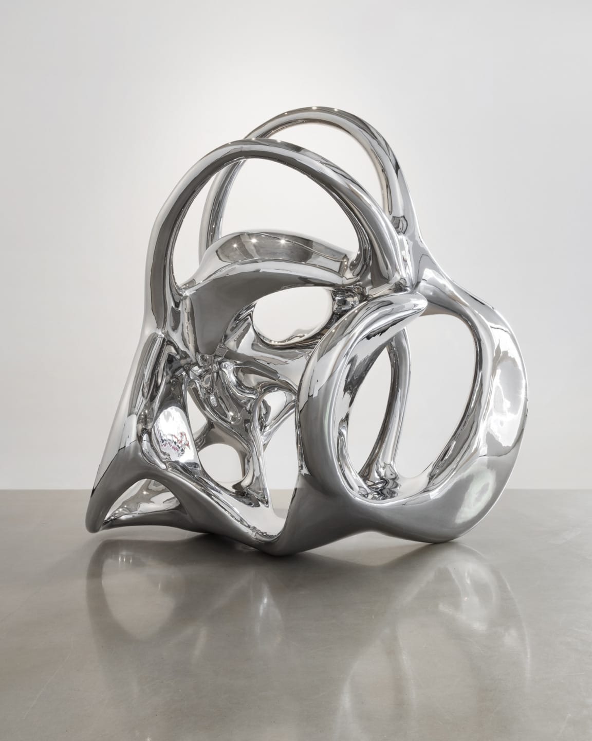 Abstract chrome sculpture with interconnected loops and smooth surfaces on a reflective floor against a plain background.