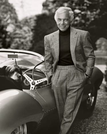 Man in a suit leaning on a classic car with a dog in the passenger seat, outdoors.