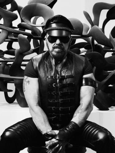 Man in leather outfit and sunglasses sitting in front of abstract sculpture, black and white photo.