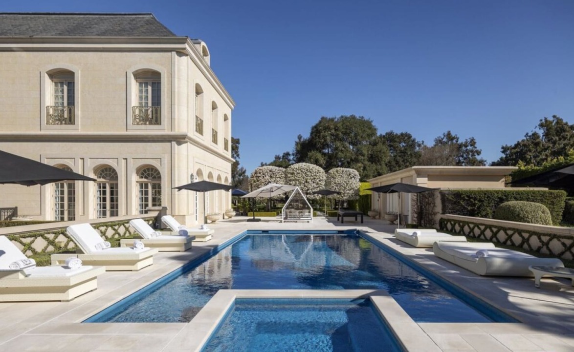 Luxurious villa with a large swimming pool, lounge chairs, and umbrellas under a clear blue sky.