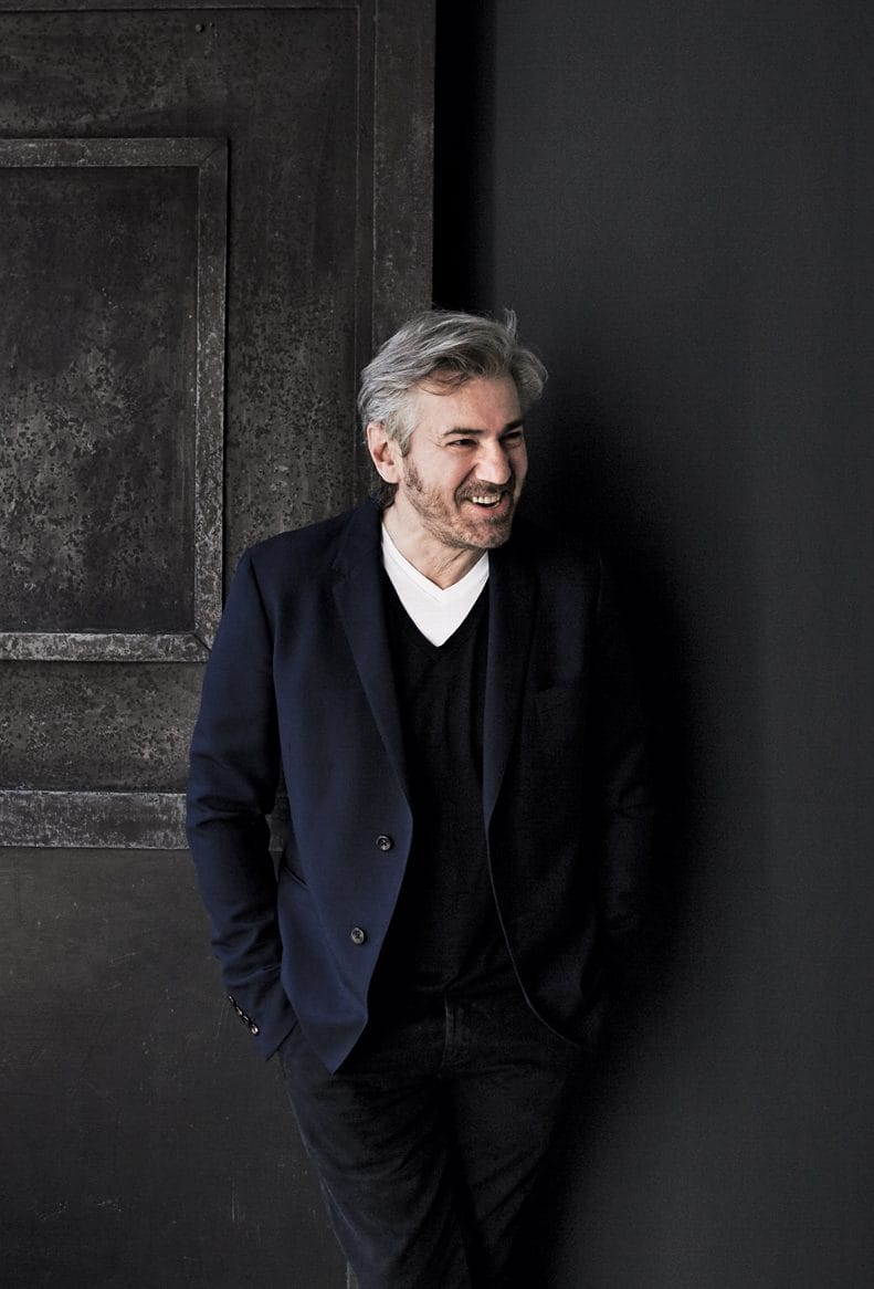 Man in a dark suit smiling, standing against a textured dark background.