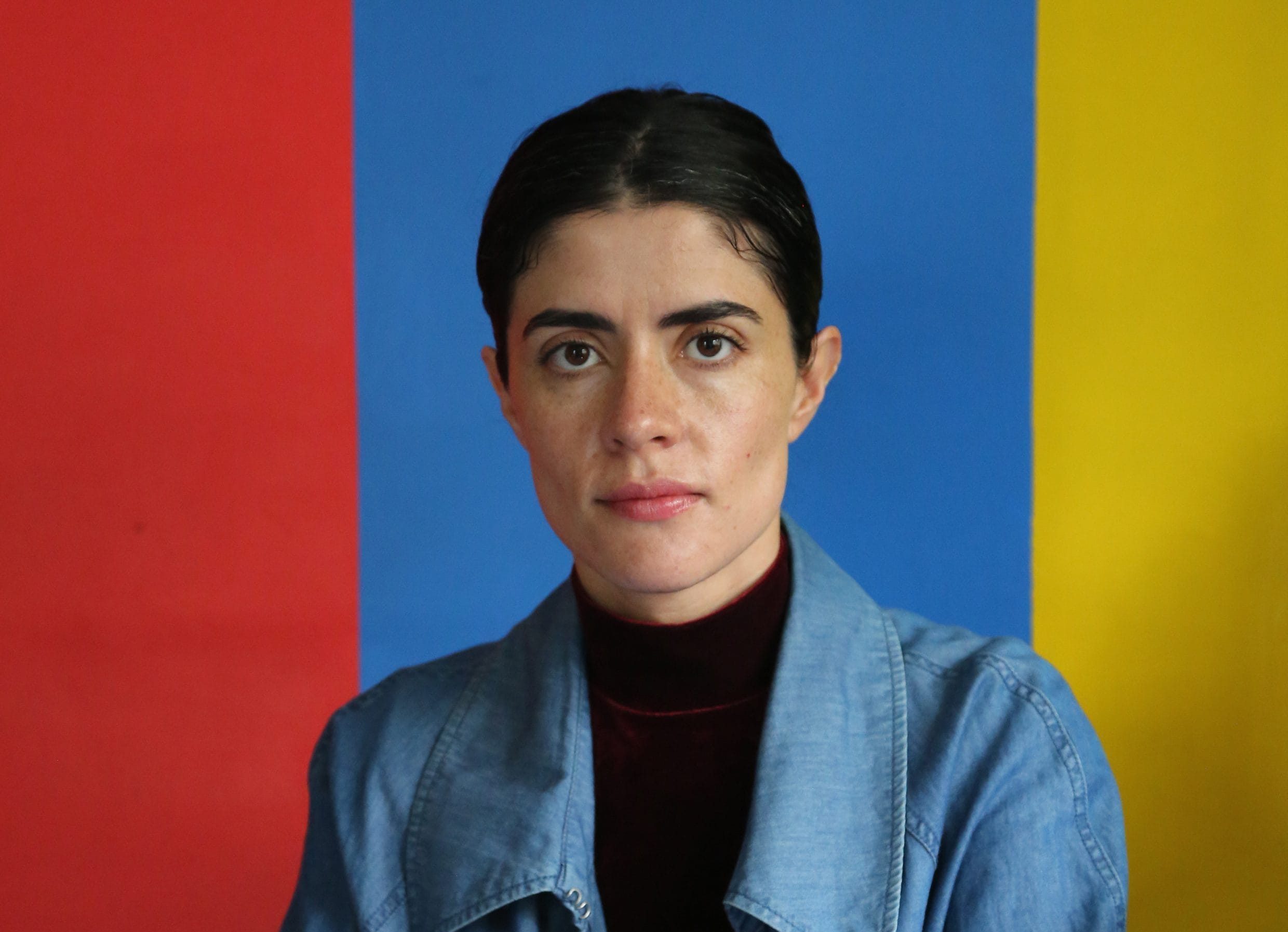 Person with short dark hair in a blue shirt stands against a red, blue, and yellow color-blocked background.