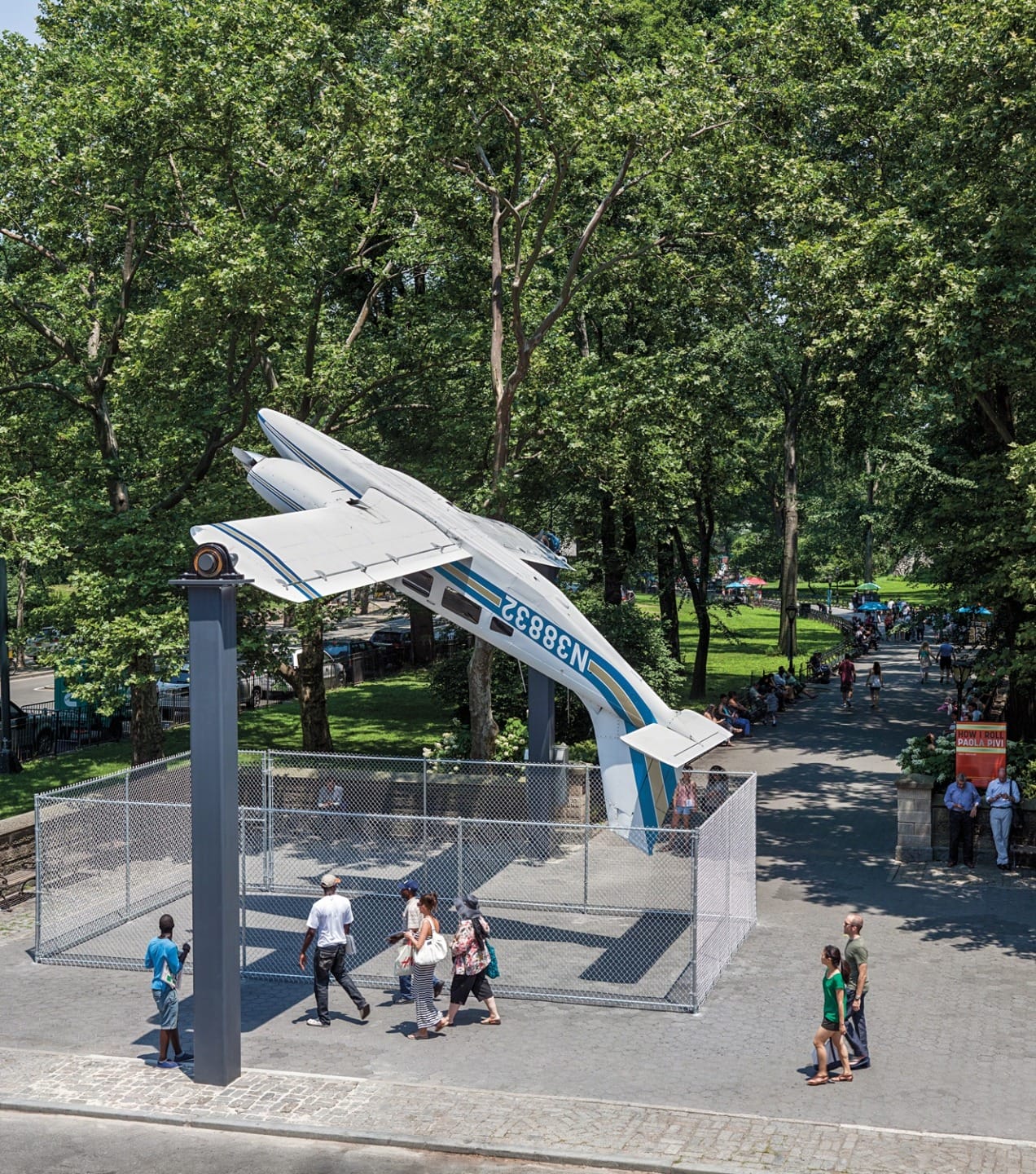 Art installation of an upside-down airplane in a park surrounded by trees, with people walking nearby.
