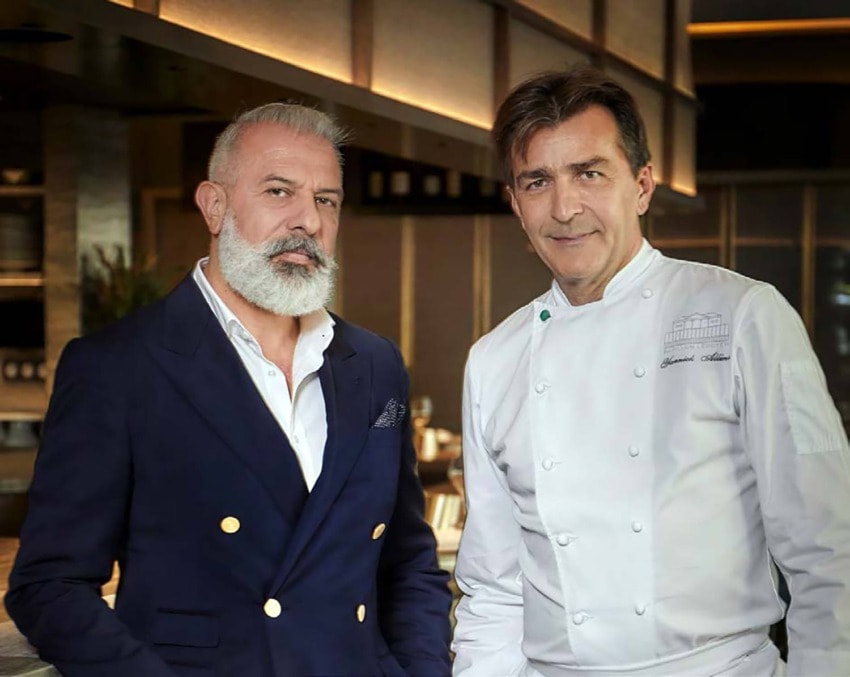 Two men in restaurant setting, one in a navy blazer with a beard and the other in a chef's coat, posing together.