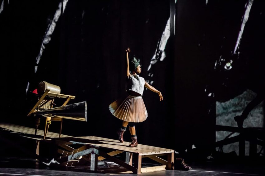 Dancer performing on stage with dynamic pose, wearing a pleated skirt and boots, surrounded by dramatic lighting and shadows.