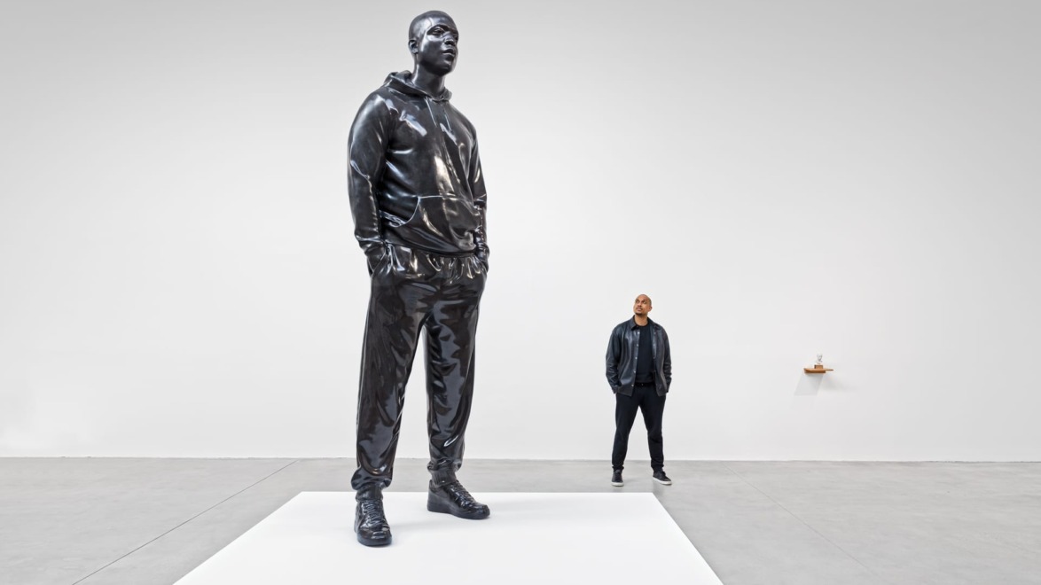 Giant black sculpture of a man in a tracksuit on a pedestal, with a person standing nearby in a gallery setting.