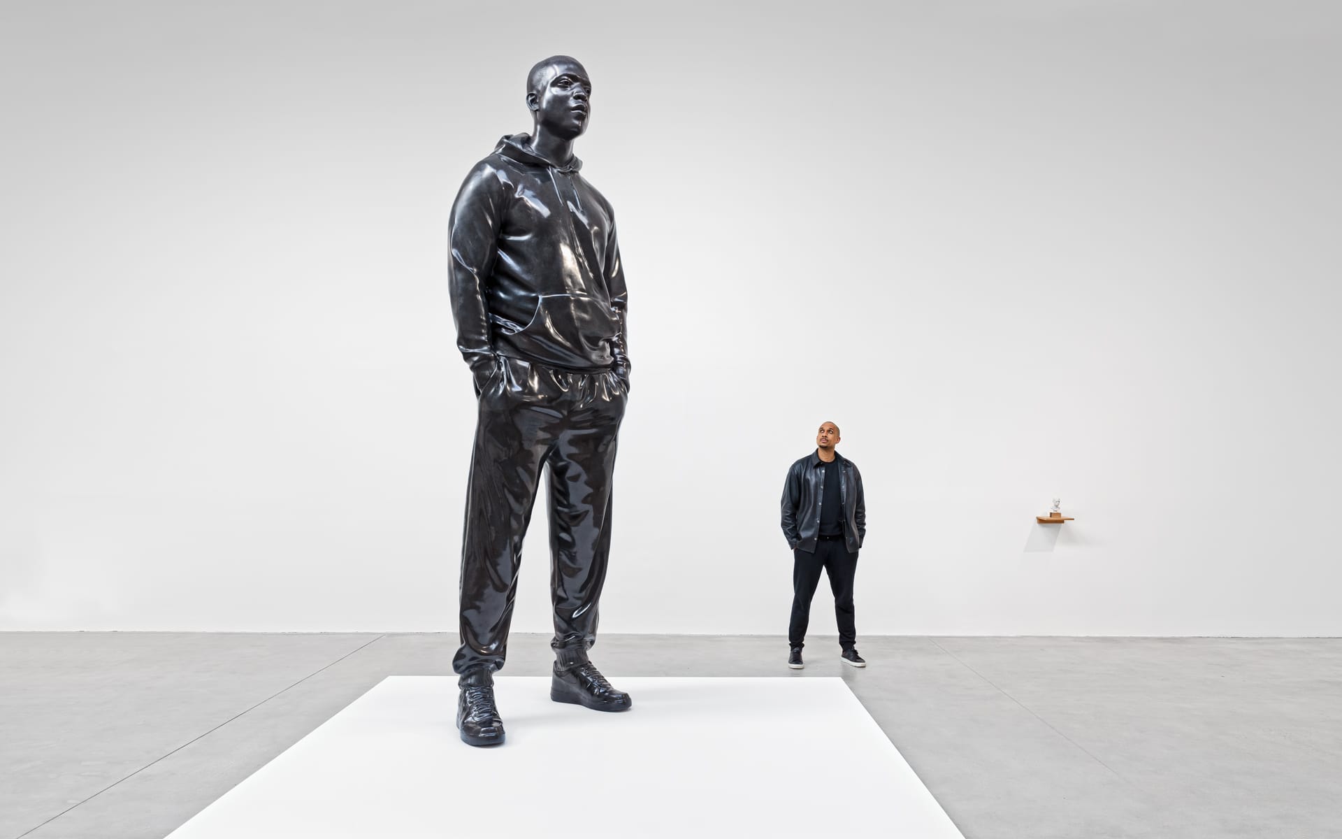Giant black sculpture of a man in a tracksuit on a pedestal, with a person standing nearby in a gallery setting.