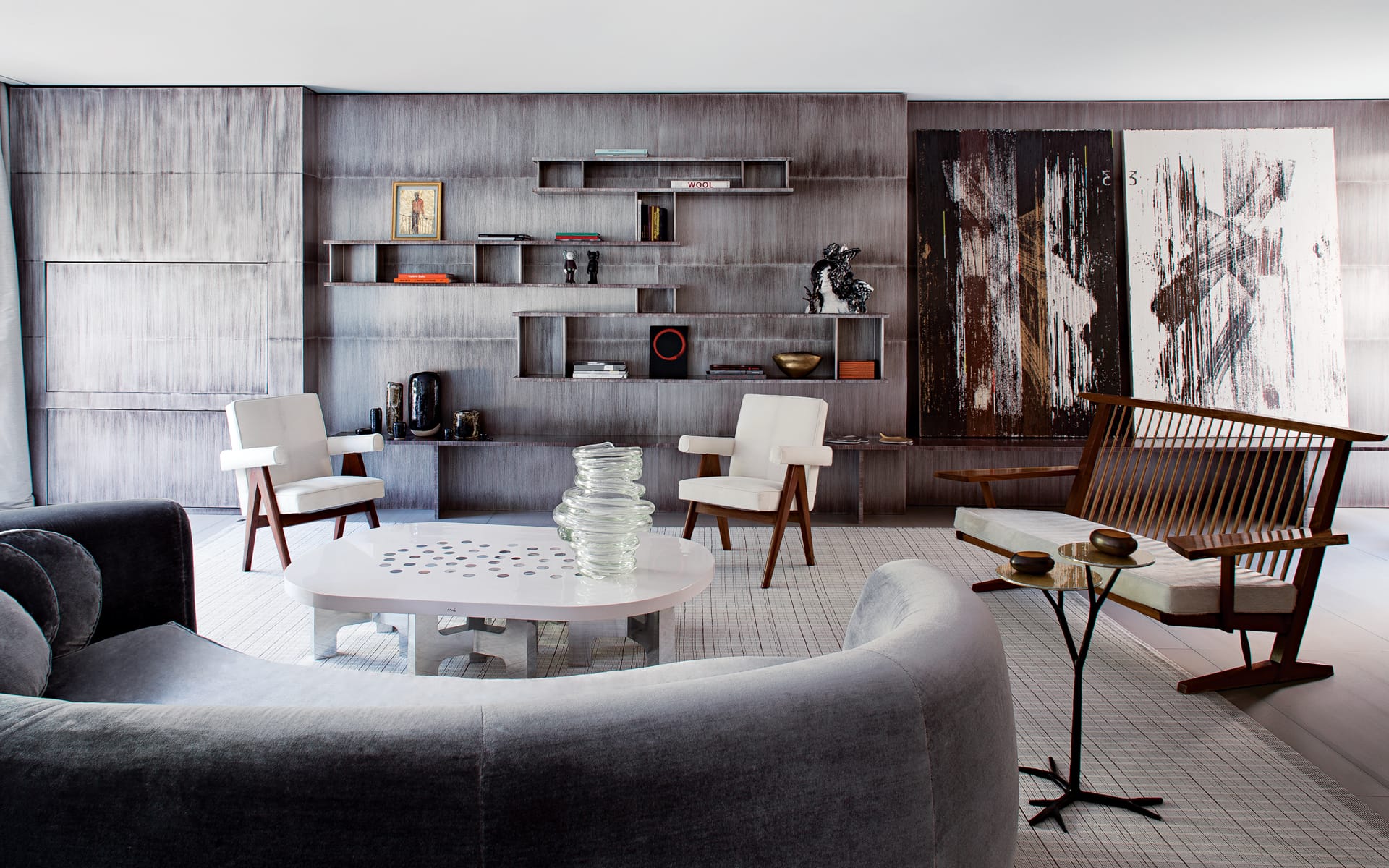 Modern living room with abstract art, grey sofa, white chairs, wooden shelves, and unique coffee table décor.