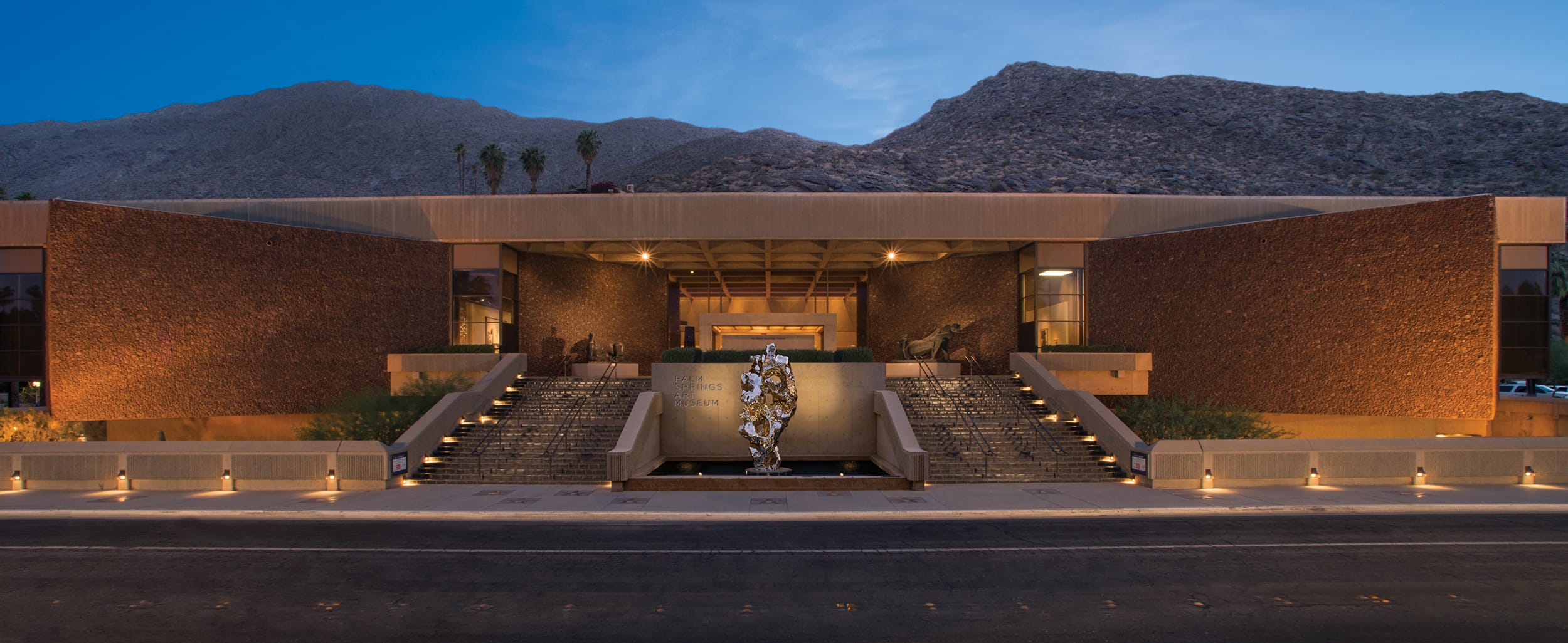 Modern building with wide steps and a large metallic sculpture at the entrance, set against a mountain backdrop at dusk.