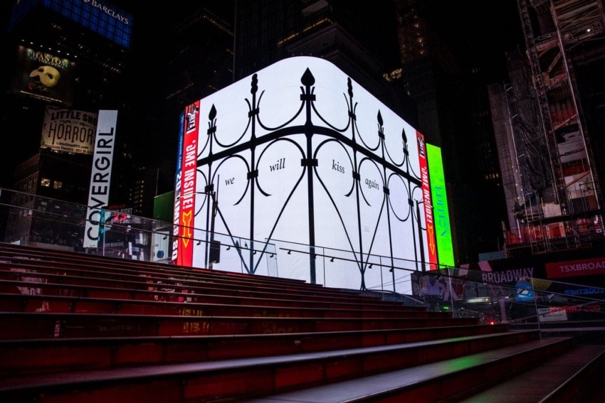 Times Square digital art display with intricate fence design and text "we will kiss again" illuminated at night.
