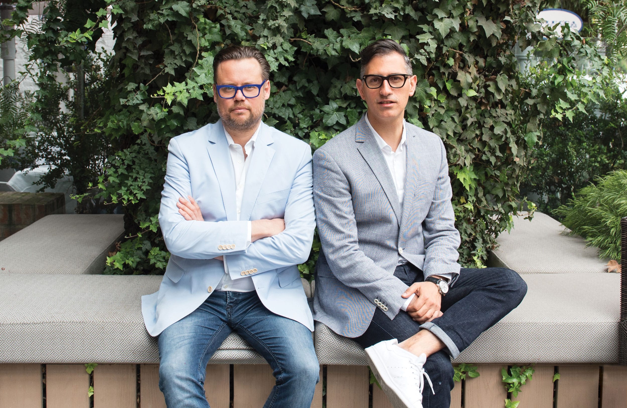 Two people sitting on a bench outdoors, wearing blazers and jeans, surrounded by greenery.