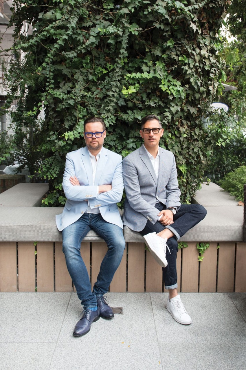 Two men in blazers sitting on a bench in front of lush ivy-covered wall outdoors.