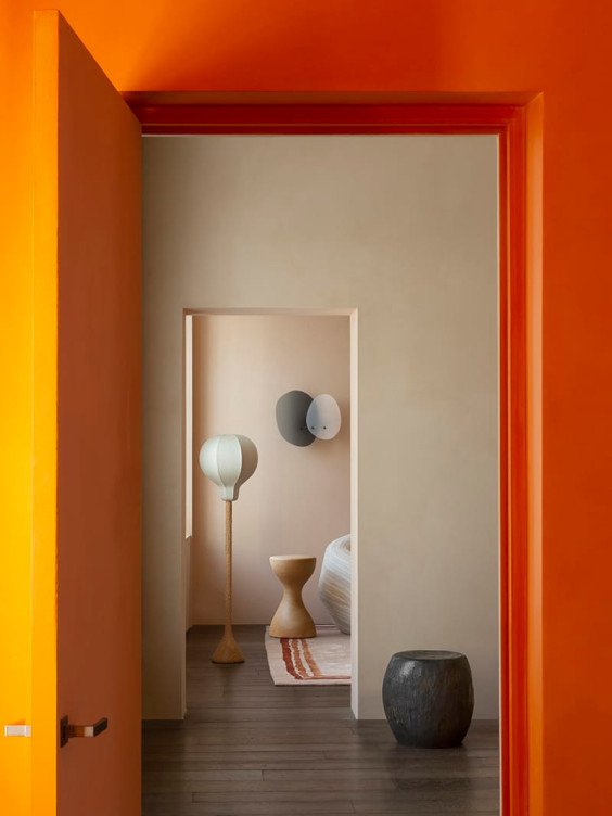 View through an open orange door into a modern room with a unique lamp, stool, and artistic decor.