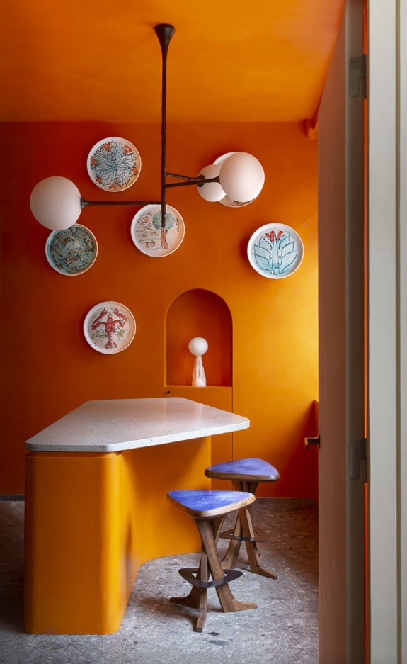 Bright orange interior with a unique countertop, decorative plates on the wall, and two wooden stools with blue seats.