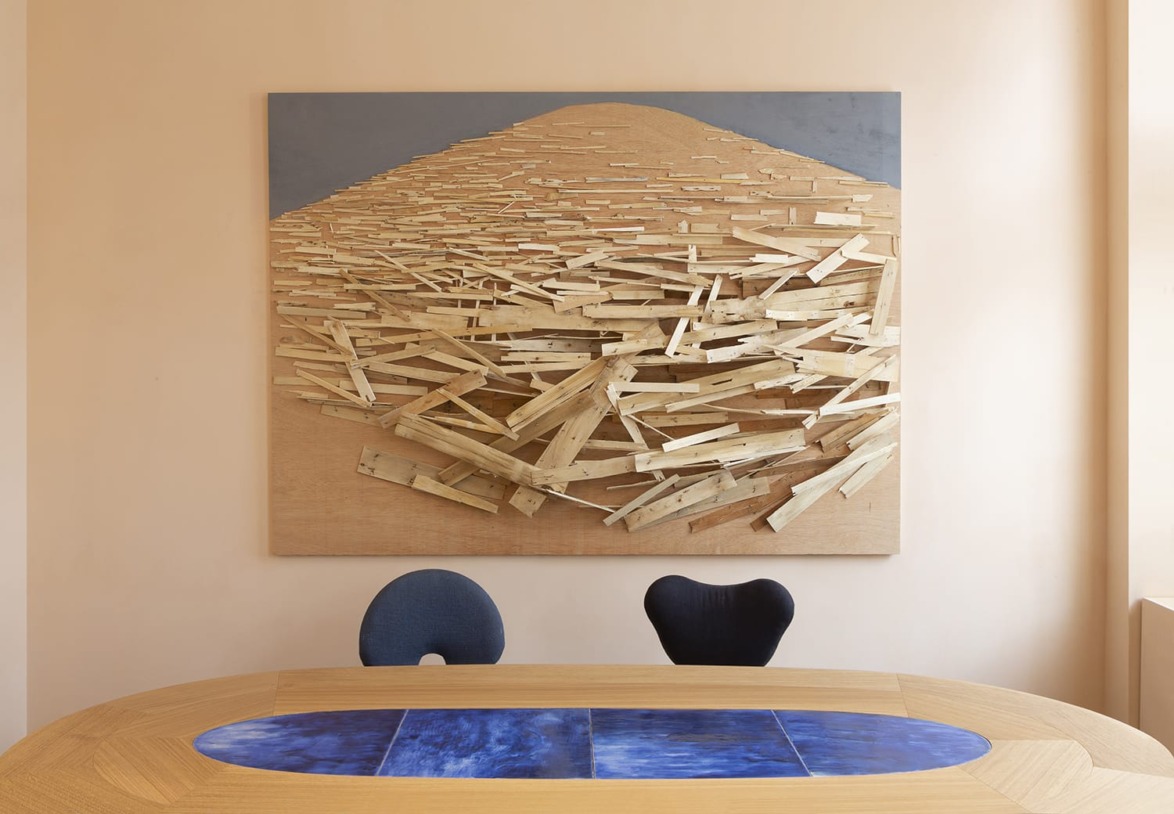 Abstract wooden art piece on the wall behind modern chairs and a wooden table with a blue center in a minimalist room.