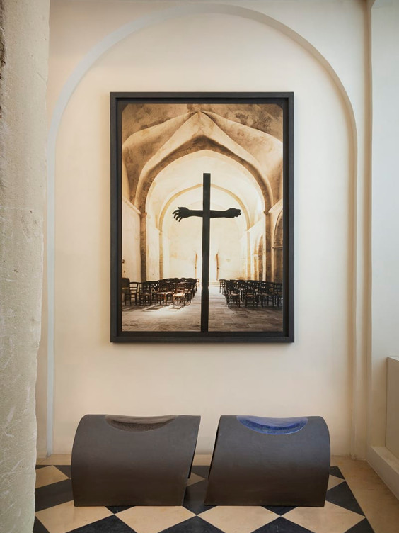Large framed cross artwork in arched room, with two modern benches and checkered floor tiles in foreground.