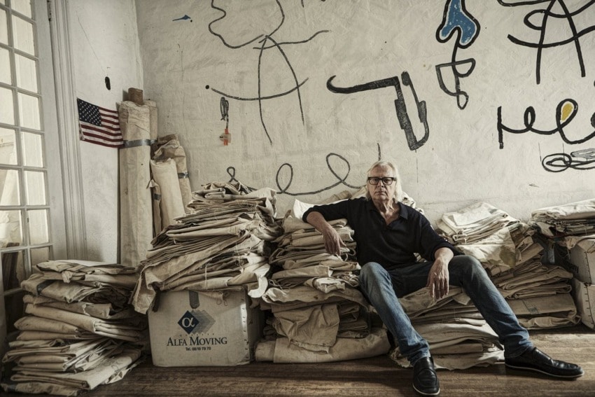 Man sitting among stacked canvases in an art studio with abstract wall sketches and an American flag.