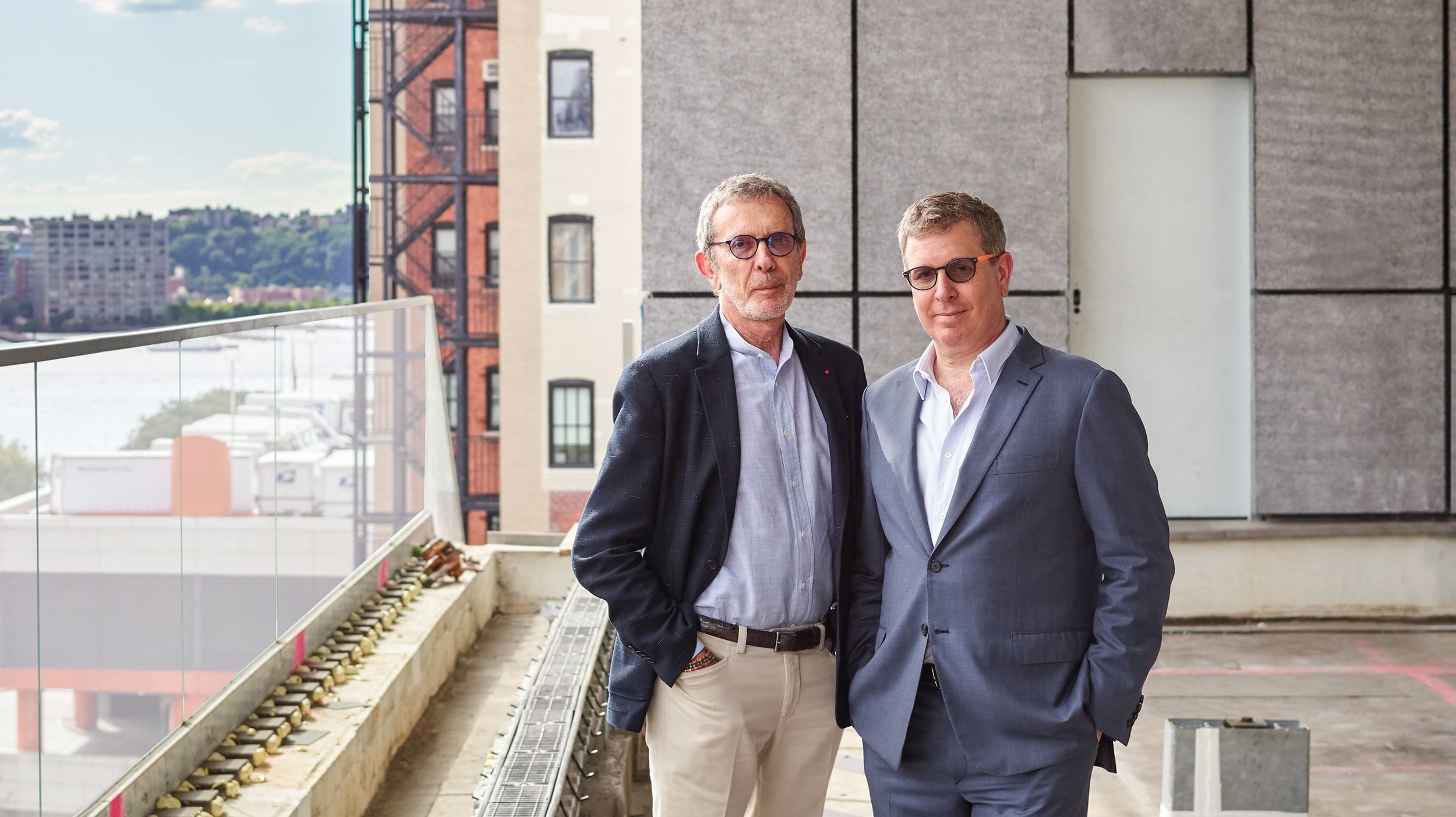 Two men in suits stand on a balcony with an urban backdrop, featuring buildings and a body of water.