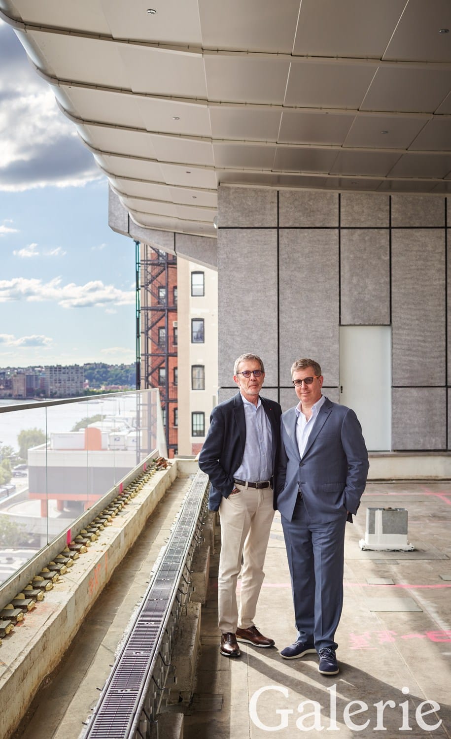 Two men in suits standing on a building balcony under construction with a cityscape view in the background.