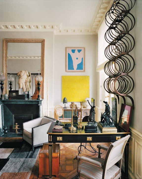 Elegant home office with ornate desk, sculptures, modern art, and large window with spiral iron decor blocking sunlight.