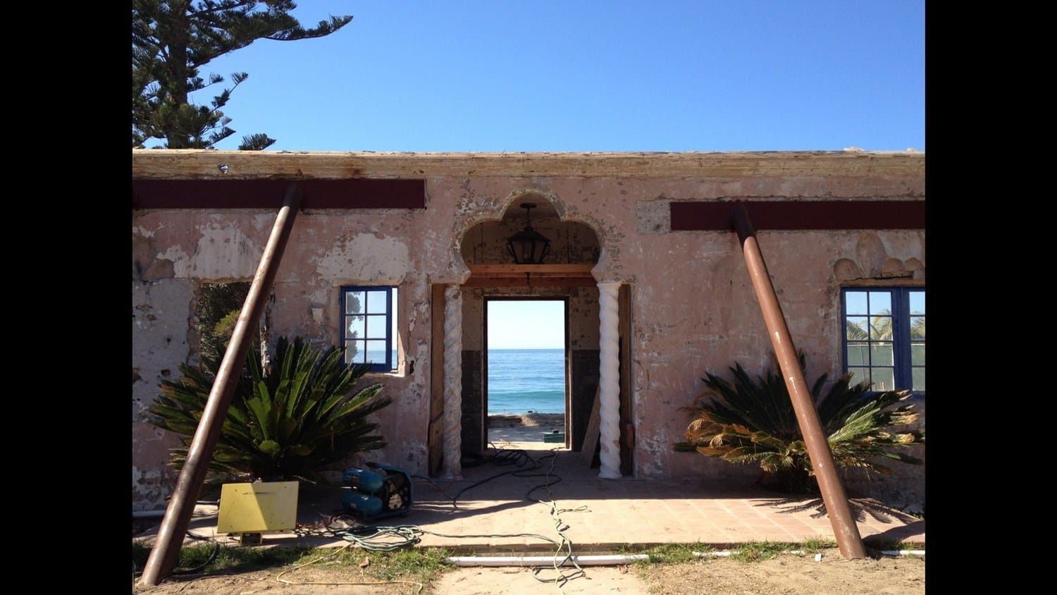 Old building facade with wooden supports, open doorway framing a view of the ocean, and clear blue sky above.