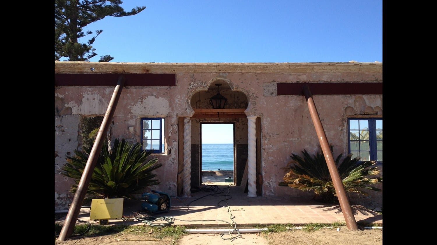 Old building facade with wooden supports, open doorway framing a view of the ocean, and clear blue sky above.
