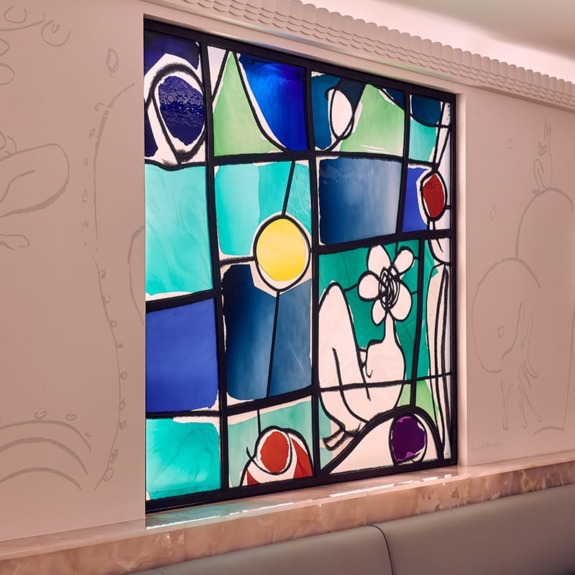 Colorful stained glass window depicting abstract shapes and a reclining figure with a flower in a bright indoor setting.