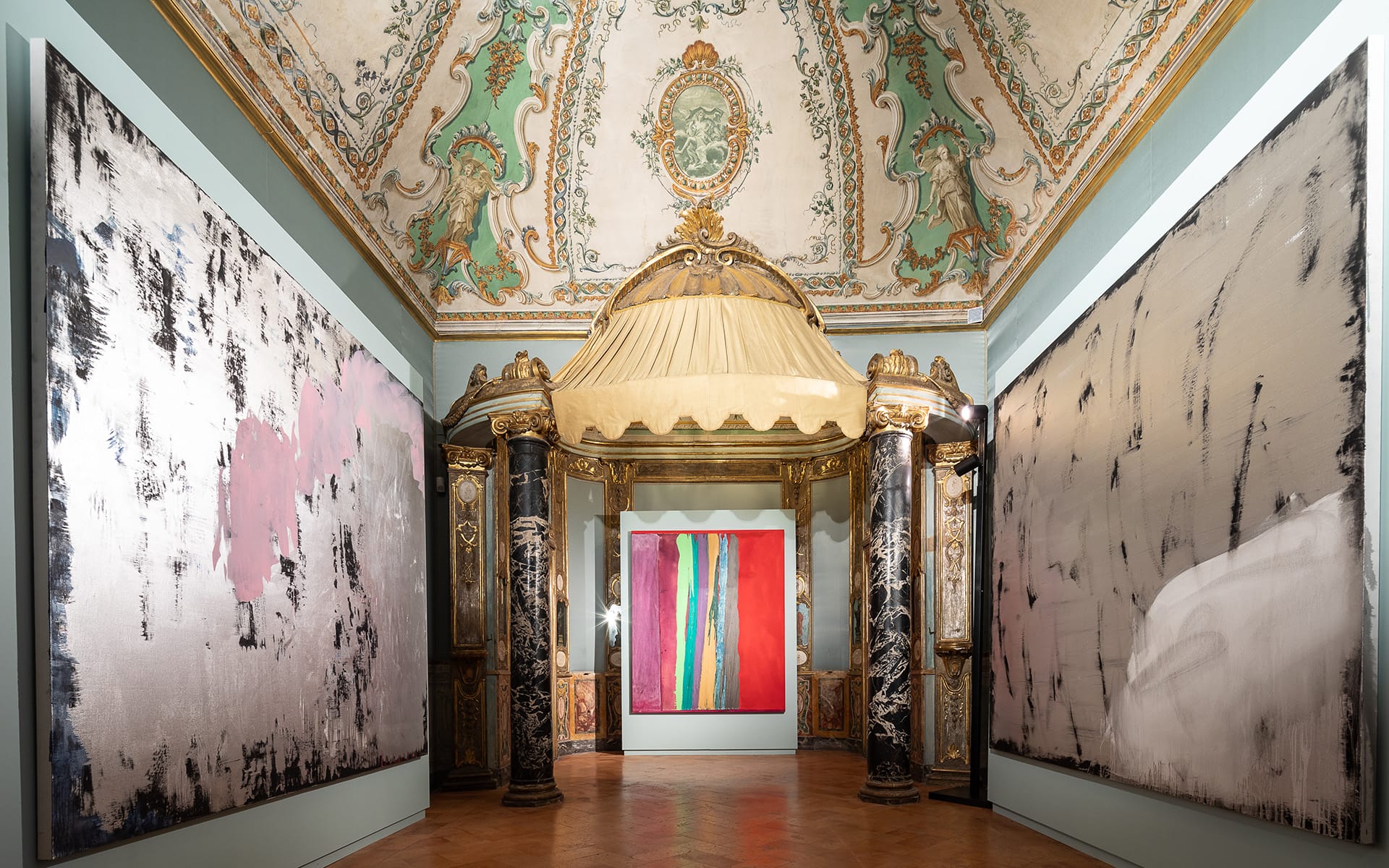 Ornate gallery with large abstract paintings on walls, elaborate ceiling design, and decorative columns surrounding a canopy.