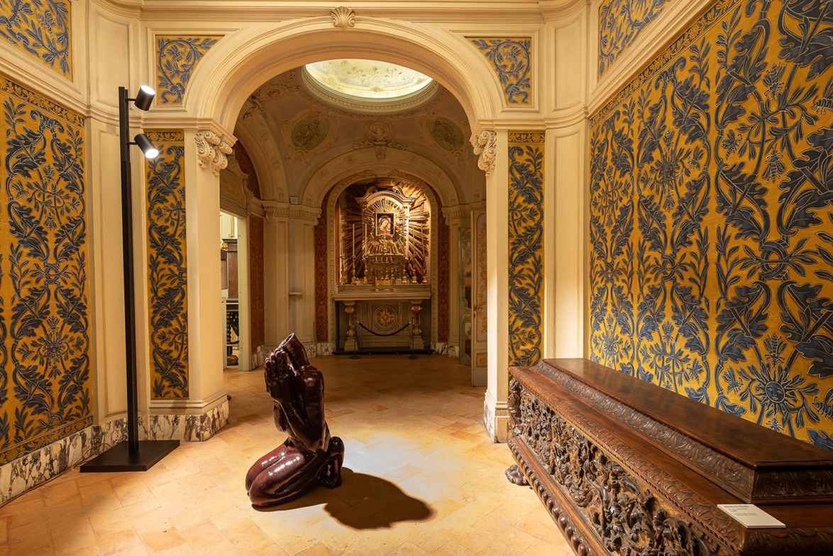 Ornate room with a statue of a kneeling figure, detailed wallpaper, carved wooden furniture, and an illuminated altar in the background.