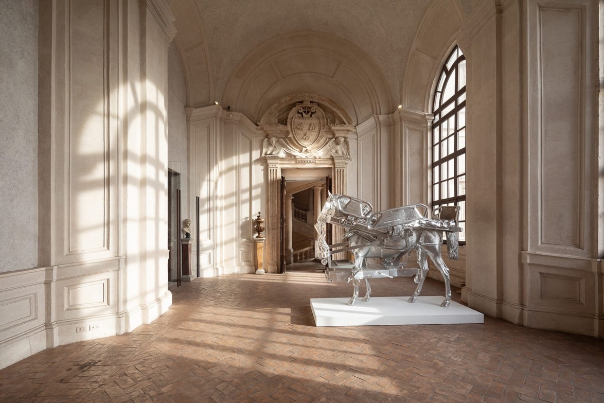 A large metal horse sculpture displayed in an ornate, sunlit hallway with high ceilings and decorative archways.